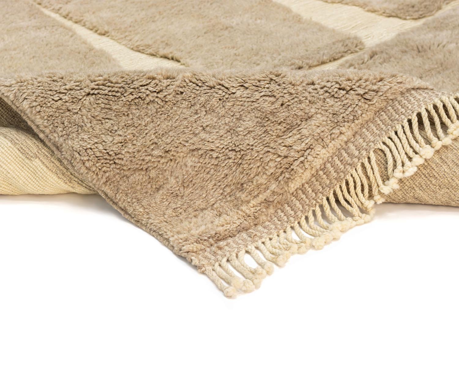 This beige, hand-knotted rug with fringed edges is artfully folded at the corner, revealing a lighter underside. Perfect for adding an elegant touch to your space, it exemplifies luxury handmade home decor.