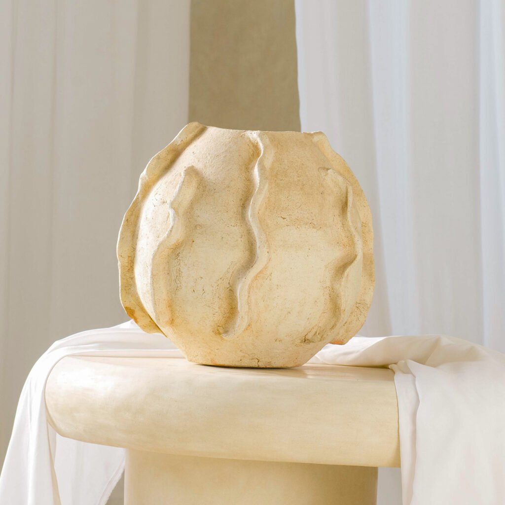 A textured, beige ceramic vase with wavy vertical ridges sits on a round pedestal, elegantly draped in a soft white fabric.