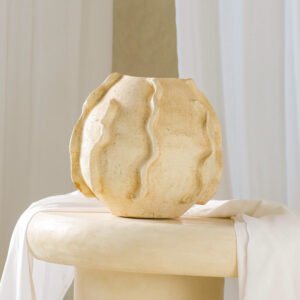 A textured, beige ceramic vase with wavy vertical ridges sits on a round pedestal, elegantly draped in a soft white fabric.