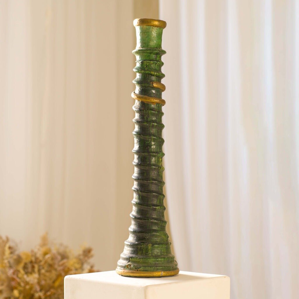 A tall, green, spiral-patterned candle holder sits elegantly on a white pedestal, with a softly blurred background of curtains and dried flowers accentuating its charm.