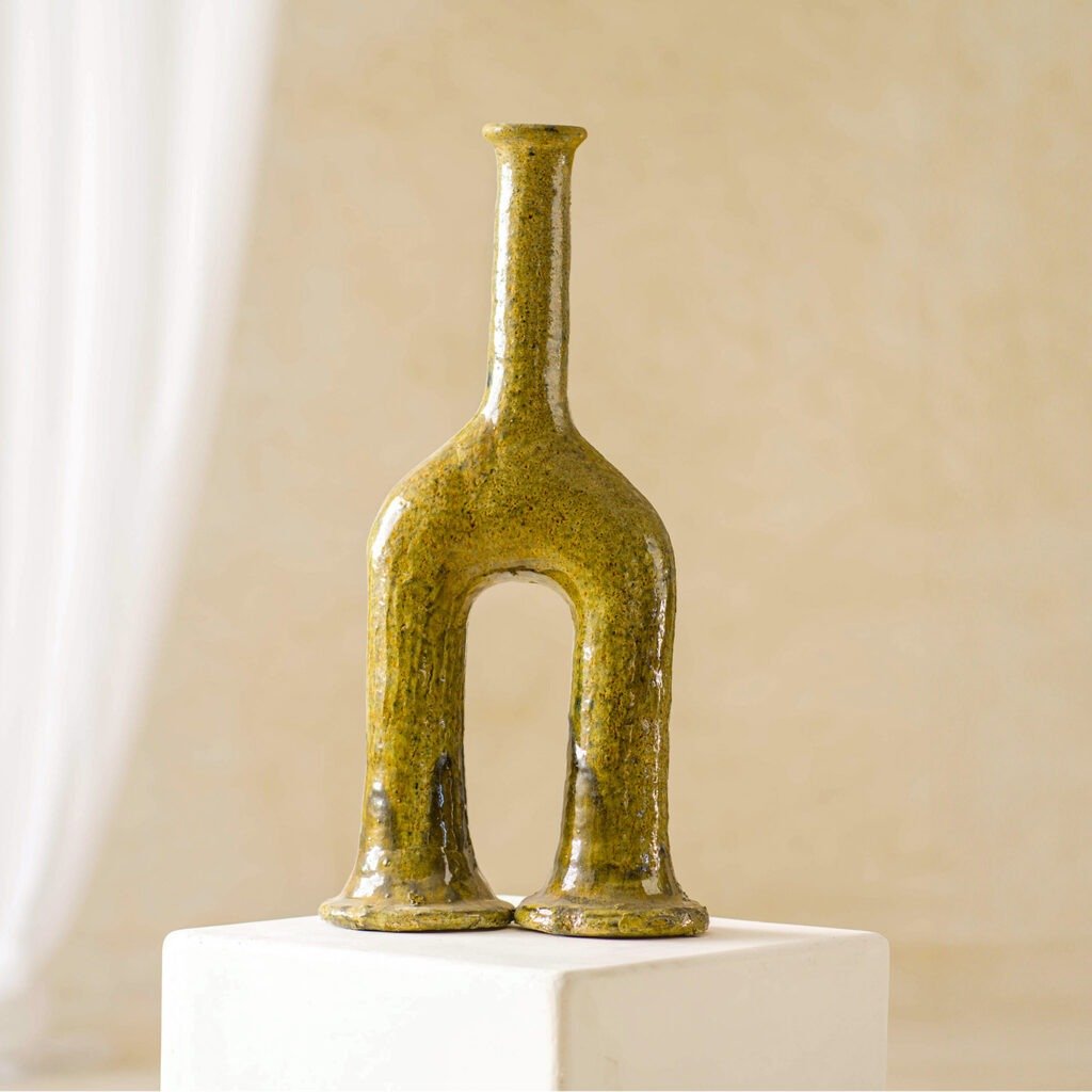 This ceramic vase features a unique double-footed design in greenish tones, elegantly displayed on a white pedestal against a soft-focus beige background. Its artistic flair makes it an eye-catching centerpiece.