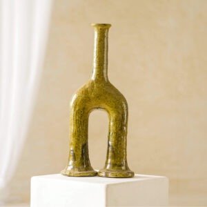 This ceramic vase features a unique double-footed design in greenish tones, elegantly displayed on a white pedestal against a soft-focus beige background. Its artistic flair makes it an eye-catching centerpiece.