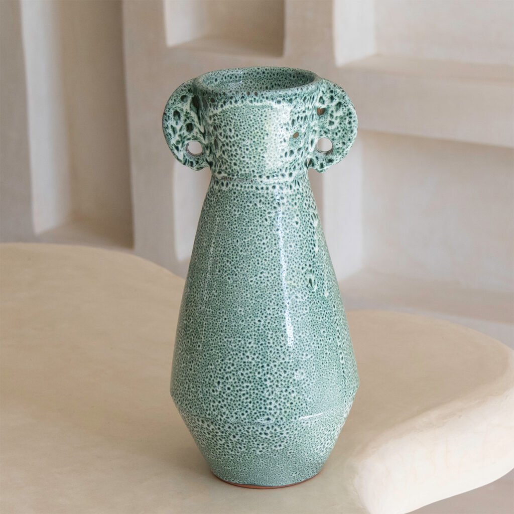 A green ceramic vase with a textured surface and two small handles sits elegantly on a light-colored surface against a neutral wall, adding a touch of artistry to any space.