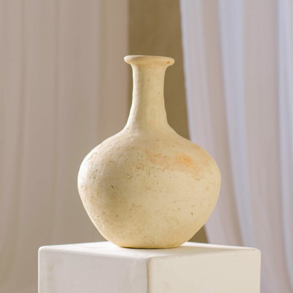 A beige ceramic vase with a narrow neck and bulbous base sits elegantly on a white platform, framed by a softly blurred background featuring light drapes.