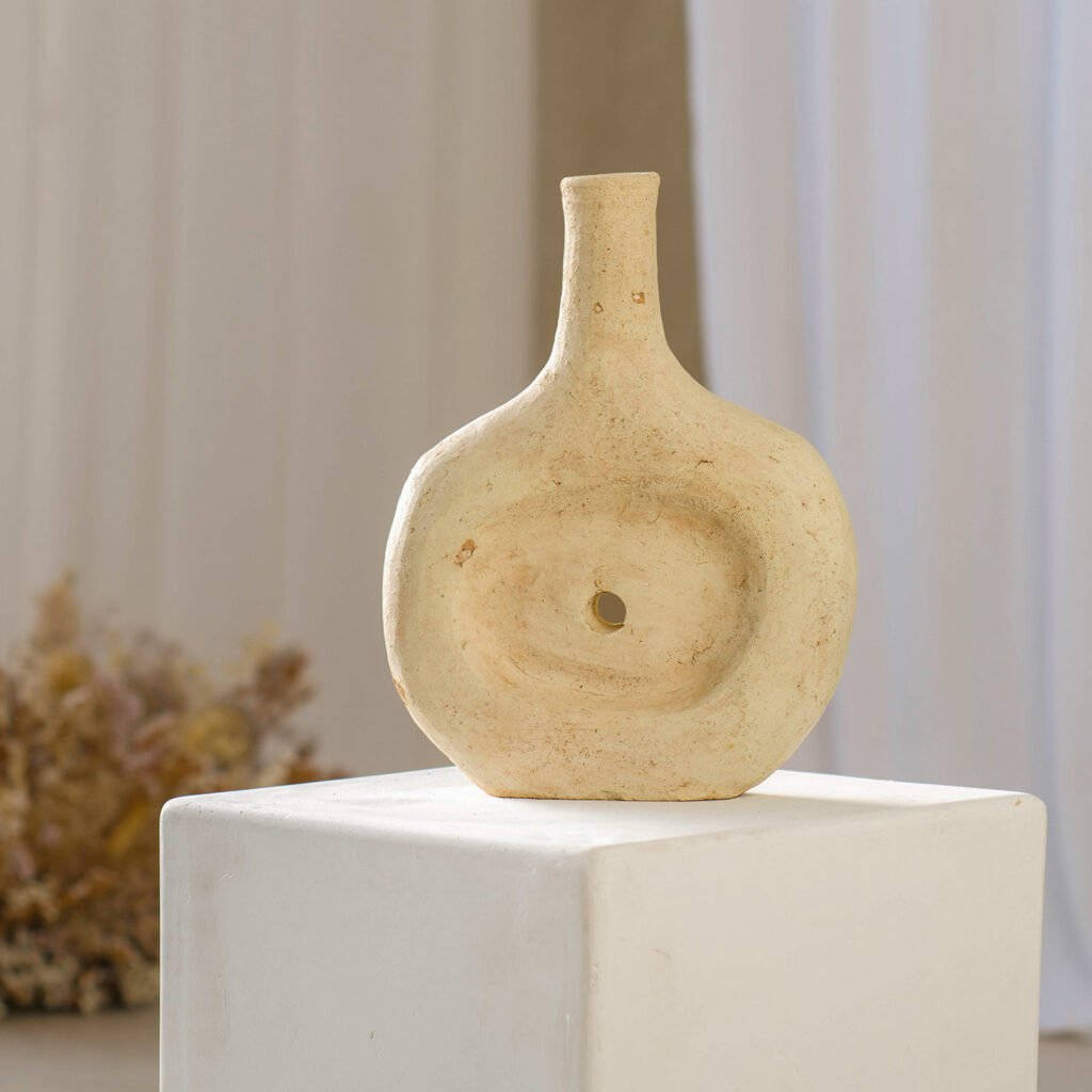 A beige, round ceramic vase featuring a small spout and central hole sits elegantly on a white pedestal. Behind it, draped curtains and soft lighting create an inviting backdrop.