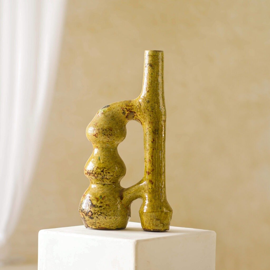 A textured, abstract ceramic vase with a unique curved design and earthy tones elegantly rests on a white pedestal against a soft beige background, showcasing its artistic charm.