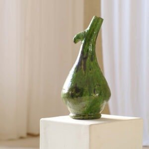 Green, abstract ceramic vase with a unique elongated, curved neck, placed on a white pedestal against a light background, exuding an artistic charm that captivates.