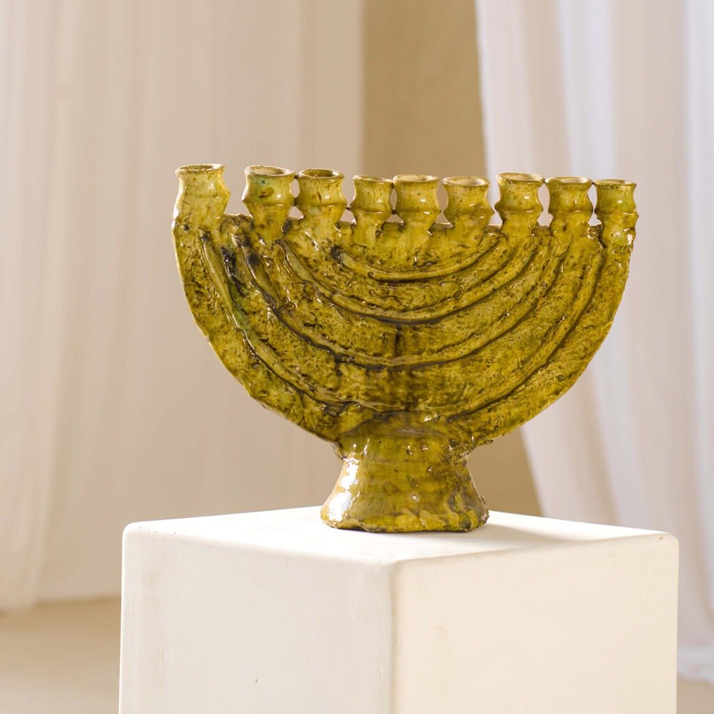 A whimsical green ceramic menorah with nine branches graces a white pedestal, set against a softly blurred backdrop.