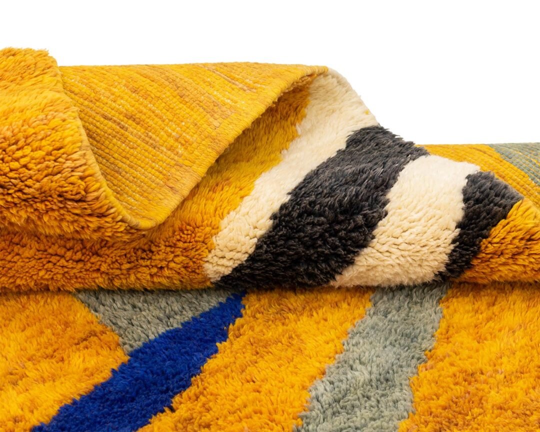 Close-up of a luxurious, colorful textured rug, showcasing yellow, blue, and gray geometric patterns. The folded edge reveals additional striped designs that exemplify exquisite craftsmanship, perfect for those seeking luxury rugs to elevate their space.