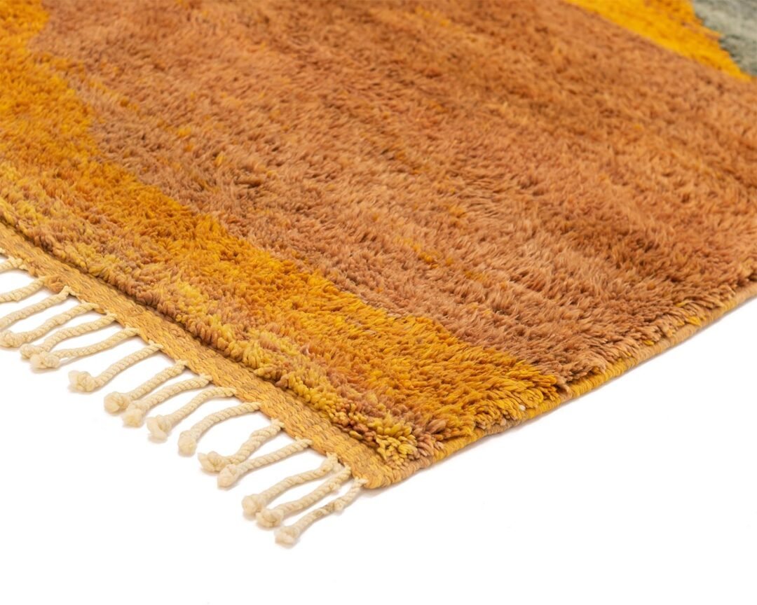 A close-up of a corner of a hand-knotted rug with fringe reveals warm-toned, textured patterns in shades of orange and yellow, epitomizing luxury rugs.