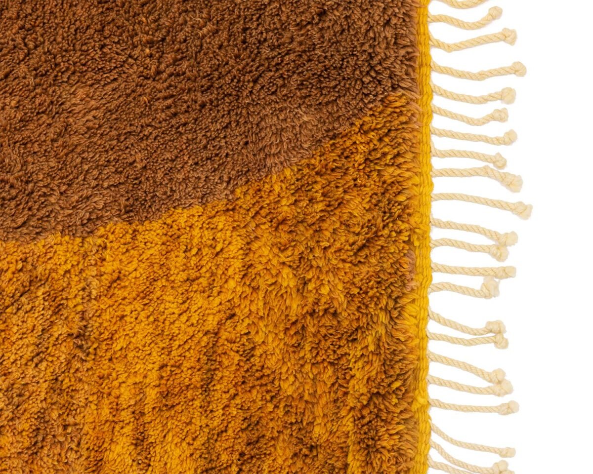 A close-up of a textured, brown and orange luxury rug with white fringes on the right edge highlights its intricate hand-knotted design.