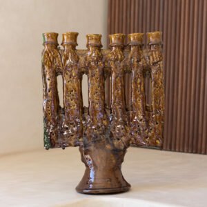 A ceramic menorah with a rustic, earthy texture and seven uneven branches is elegantly displayed on a light-colored surface with a wooden backdrop, creating an inviting ambiance.