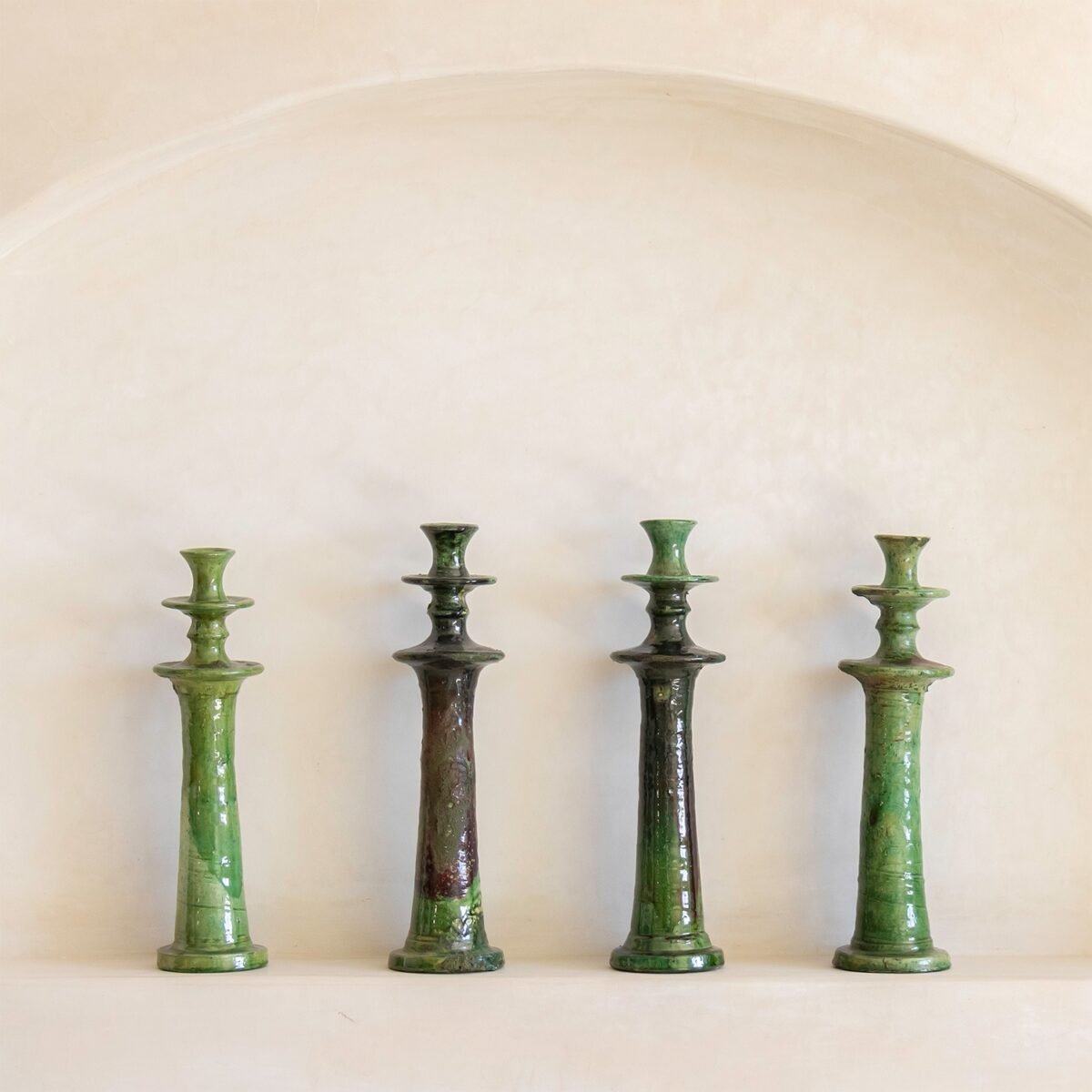 Four green ceramic candlesticks, each with unique shades and patterns, stand gracefully on a white shelf against an arched, textured beige wall. The varying greens of the candlesticks bring a refreshing vibrancy to the serene setting.