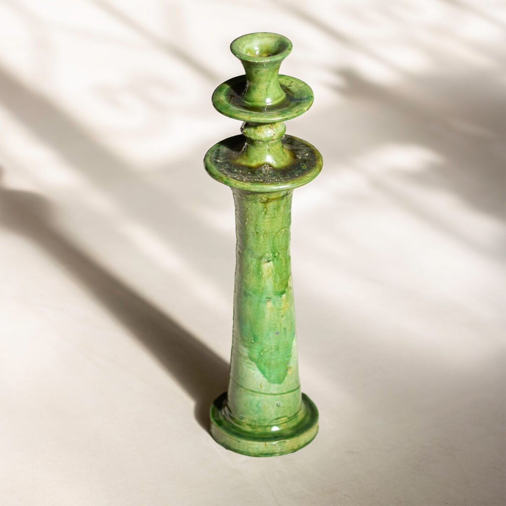 A tall, green ceramic candlestick, crafted in the style of Tamegroute pottery, stands on a light surface with shadows in the background.