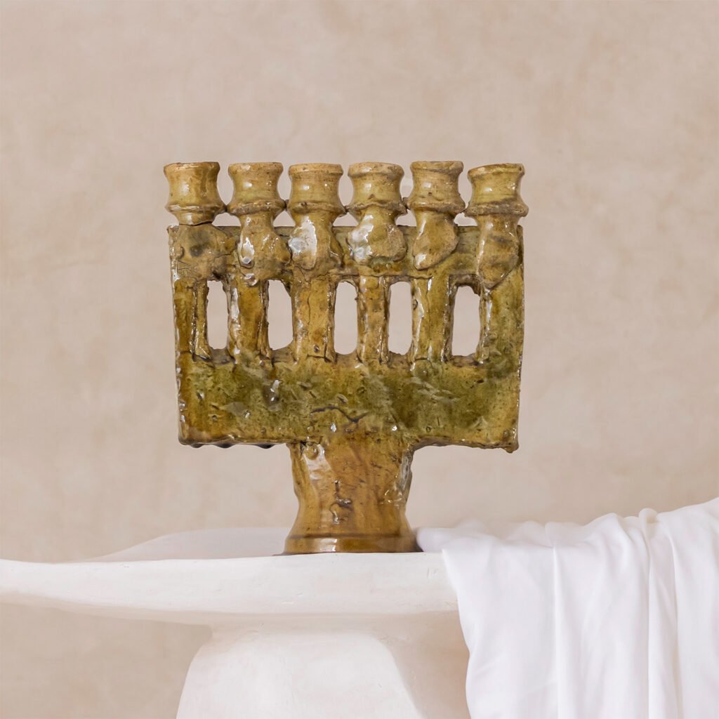 A rustic, seven-branched menorah with a textured, earthy finish stands on a draped white surface against a calming beige backdrop.
