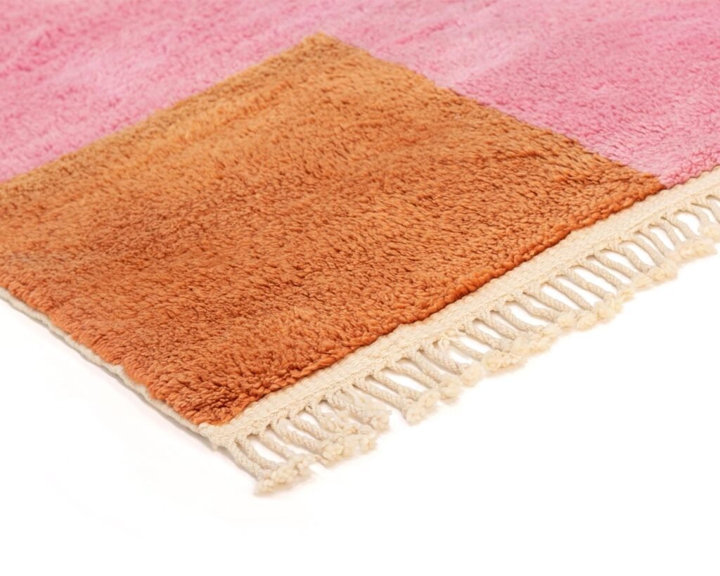 A close-up of a fringed rug corner showcasing a vibrant pink and orange square design, accented by white fringe along the edge on a simple backdrop.