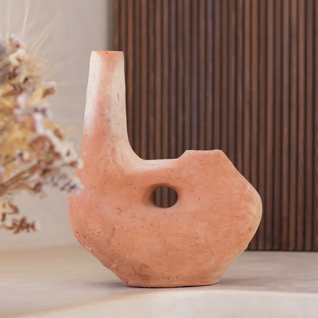 A rustic terracotta vase with a unique asymmetrical design, featuring a hole in the center, is artfully displayed against a wooden slat wall. Dried flowers gracefully peek out from the left side, adding charm and texture to this elegant setting.