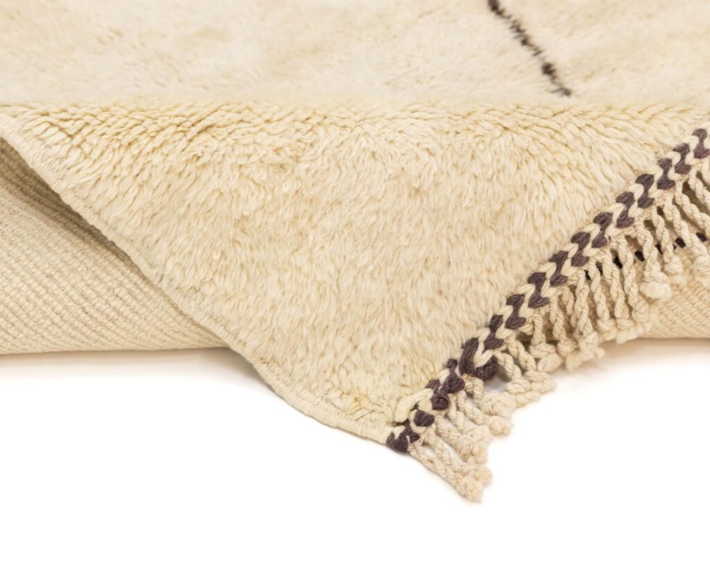 A close-up of a luxurious, textured cream-colored rug with fringed edges and a subtle brown pattern, showcasing the intricate craftsmanship typical of hand knotted rugs.