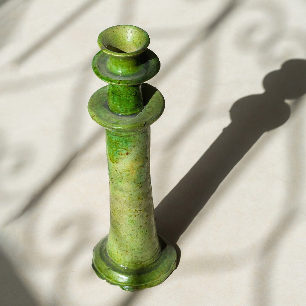 A green ceramic candlestick casts a long shadow on a light surface, creating soft, swirling patterns in the background. The elegant play of light highlights the craftsmanship and artistry of the piece.