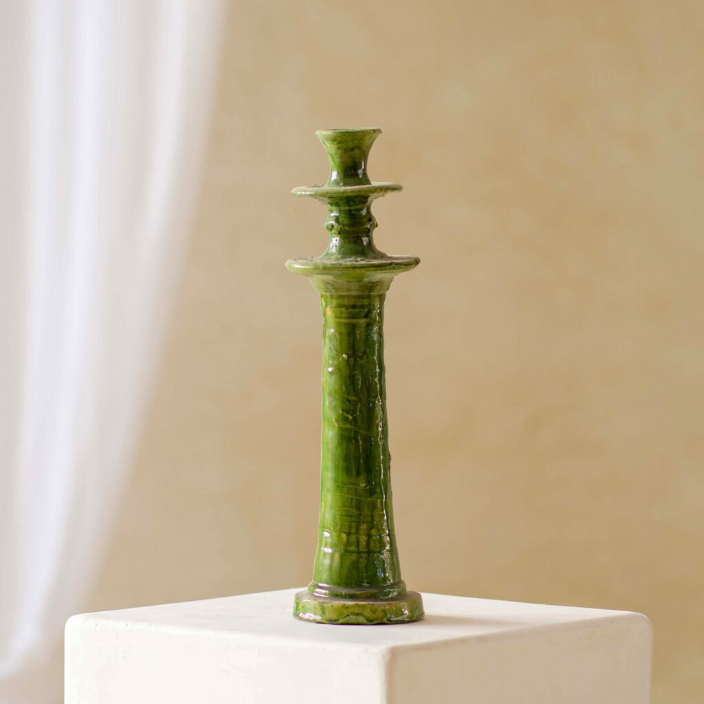 A tall green ceramic candle holder with a glossy finish stands majestically on a white pedestal against a light beige backdrop, adding elegance to any space.