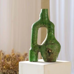 An abstract green ceramic sculpture with a curved, open design and a hollow center sits elegantly on a white pedestal. The blurred silhouette of dried flowers in the background complements the pieces artistry.