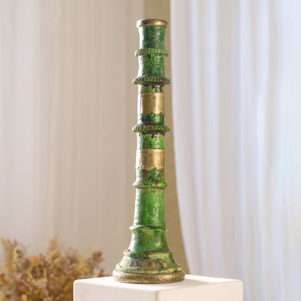 A tall, green and bronze decorative pillar with ornate details stands elegantly on a white pedestal, set against a soft background with dried plants for an artful and serene display.