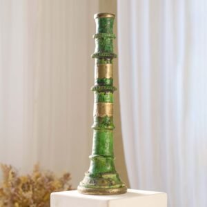 A tall, green and bronze decorative pillar with ornate details stands elegantly on a white pedestal, set against a soft background with dried plants for an artful and serene display.