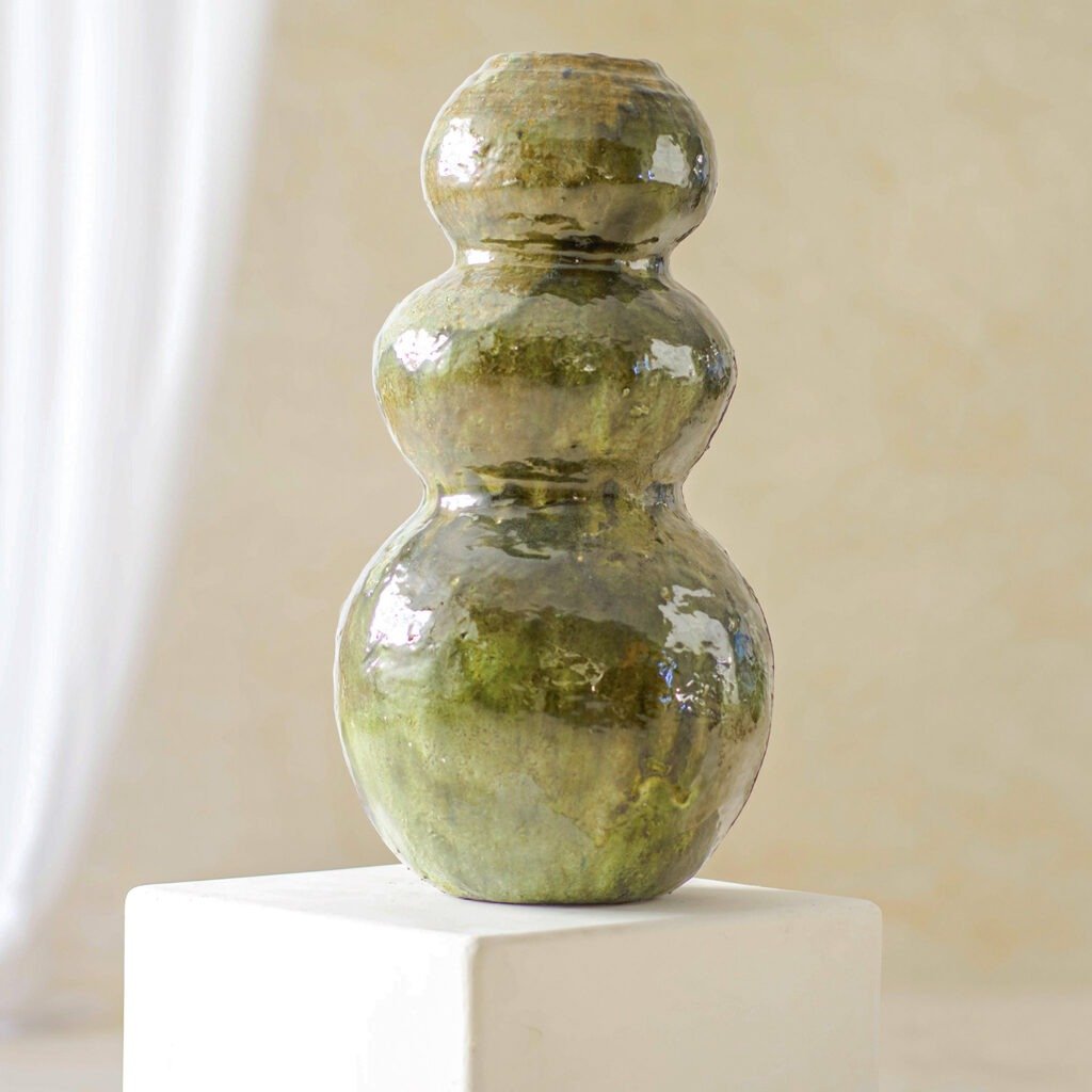 A green, glossy, triple-gourd-shaped ceramic vase elegantly stands on a white pedestal, set against a neutral background.