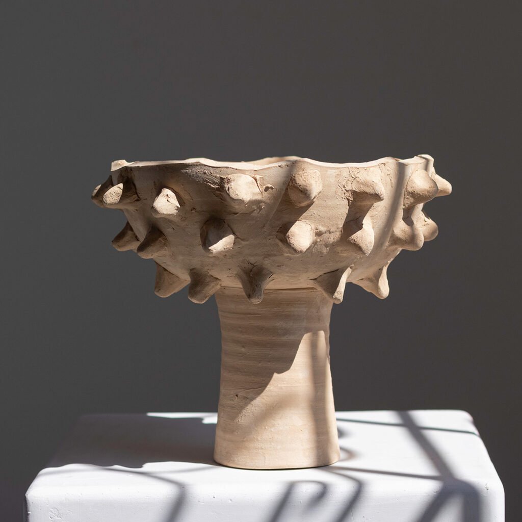 The clay sculpture stands on a pedestal, showcasing its textured, spiked surface rising from a narrow base. Dramatic lighting enhances its intricate details against the gray backdrop.