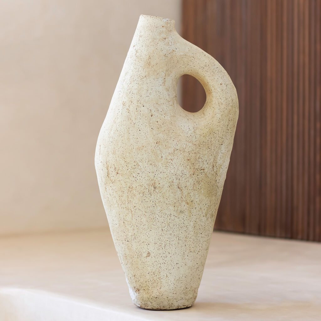 A textured, beige ceramic vase with a rounded body and an integrated handle sits gracefully on a neutral surface against a warm wooden backdrop, exuding rustic charm.