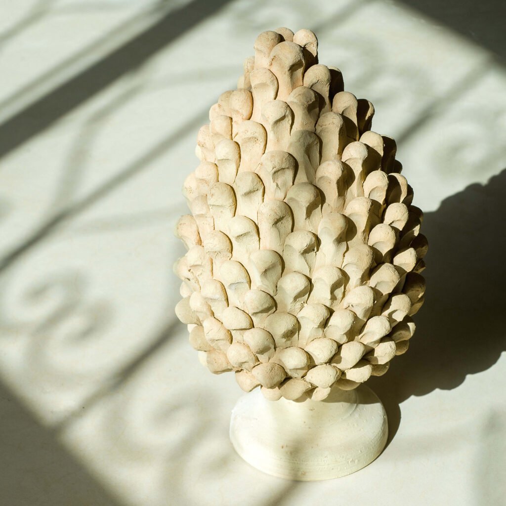 A textured ceramic sculpture resembling a pine cone stands on a light surface, with intricate shadows elegantly cast around it, bringing to mind the delicate forms found in nature.