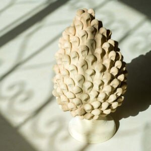 A textured ceramic sculpture resembling a pine cone stands on a light surface, with intricate shadows elegantly cast around it, bringing to mind the delicate forms found in nature.