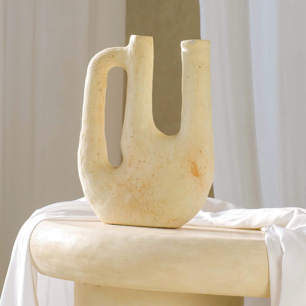 A cream-colored, abstract ceramic vase with two spouts and a handle sits gracefully on a rounded surface, complemented by the elegant texture of a luxury rug in the background.