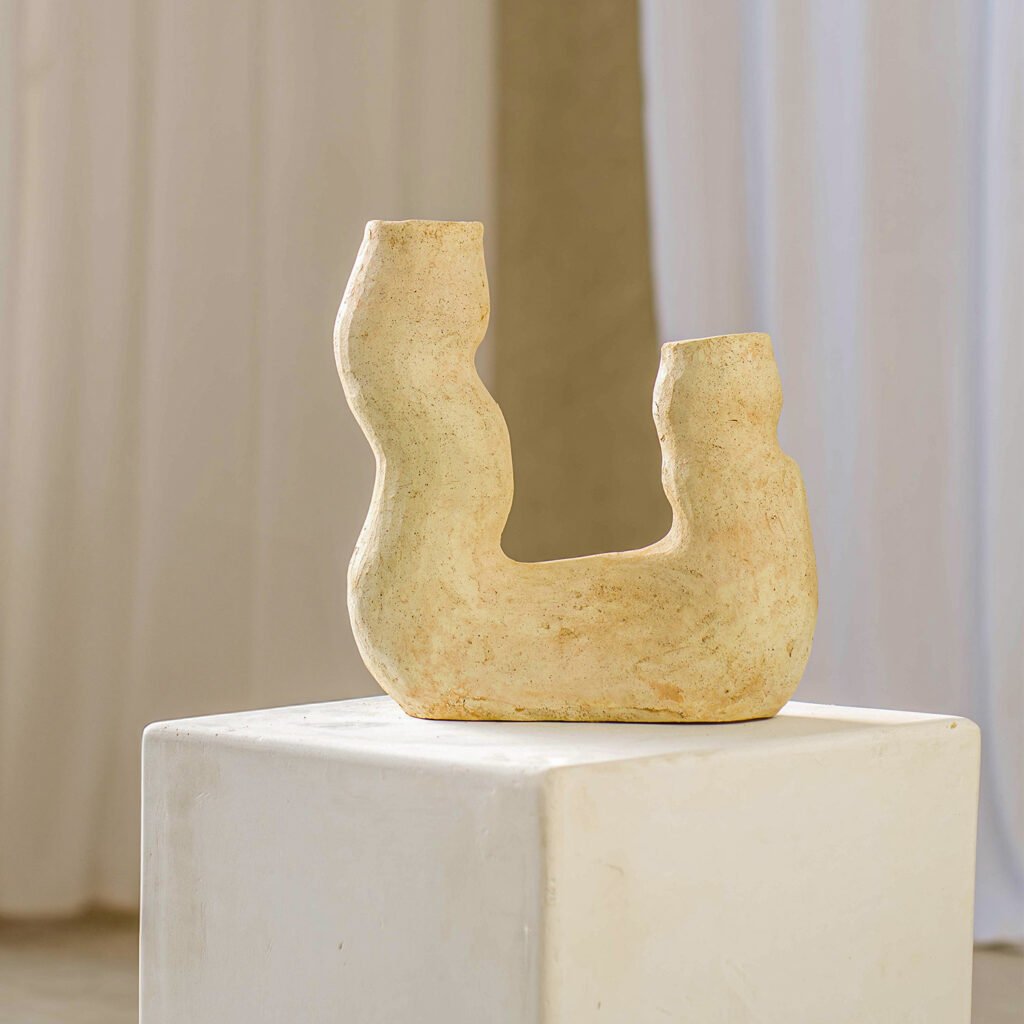 A beige, abstract clay sculpture with curved forms rests elegantly on a white pedestal, set against a backdrop of soft white drapery and a neutral wall.