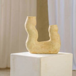 A beige, abstract clay sculpture with curved forms rests elegantly on a white pedestal, set against a backdrop of soft white drapery and a neutral wall.