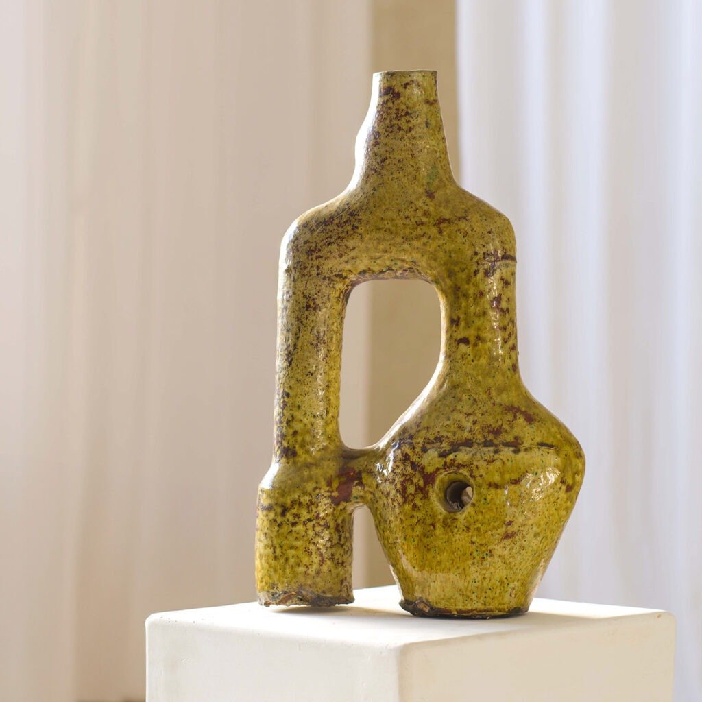 The abstract ceramic sculpture, coated in a yellowish-brown glaze, showcases geometric shapes and a hollow center. Elegantly positioned on a sleek white pedestal, its intriguing form captivates the viewers attention with its modern artistic expression.