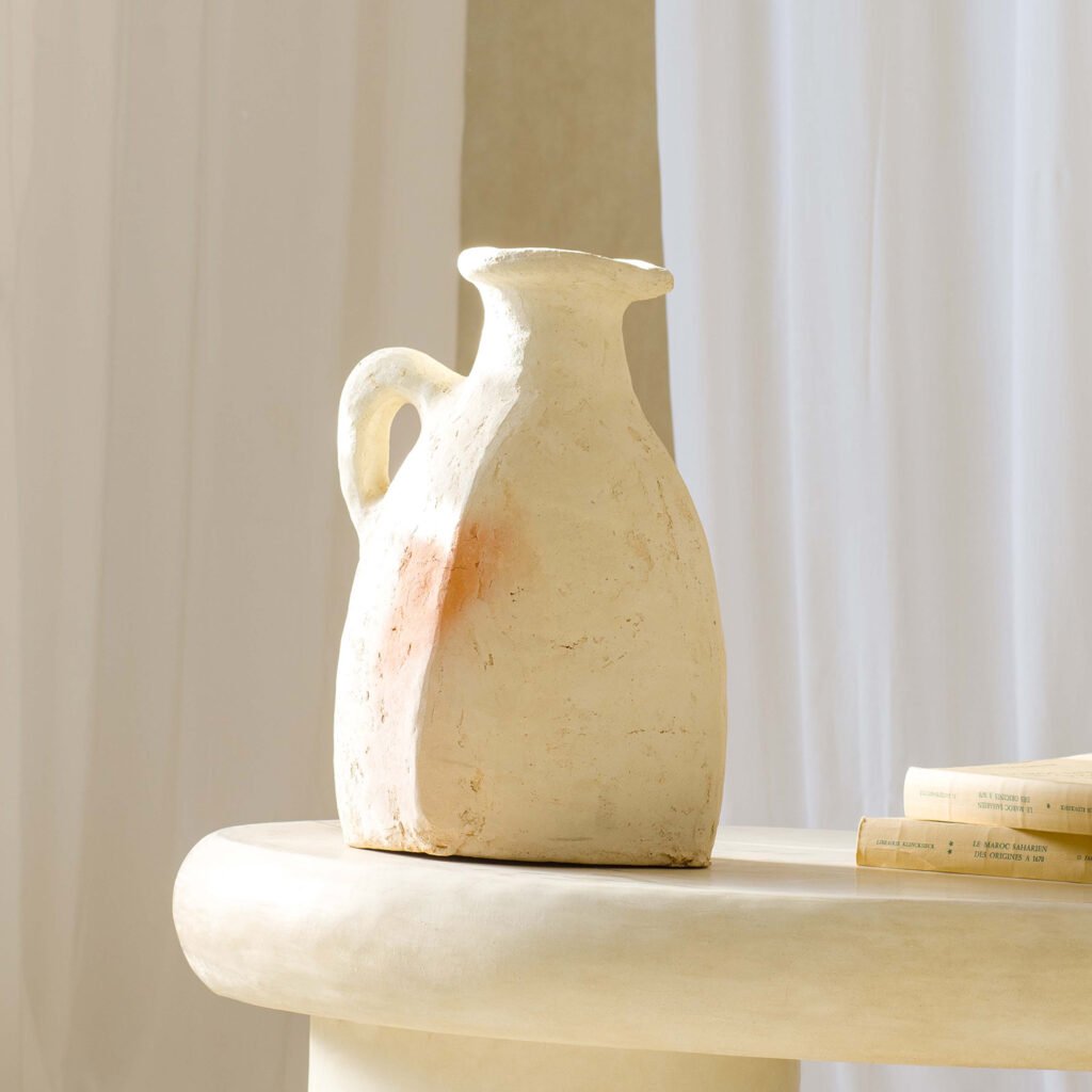 A rustic ceramic jug sits on a light-colored table beside rolled sheets of paper, illuminated by soft sunlight streaming through a sheer curtain in the background.