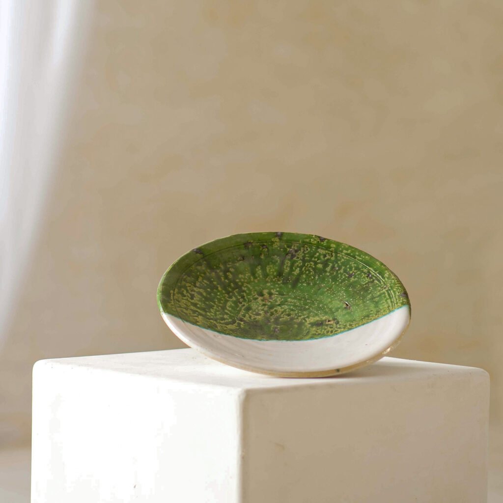 A shallow, round ceramic dish with a captivating green interior glaze delicately rests on a white pedestal, creating an elegant contrast against the neutral background.