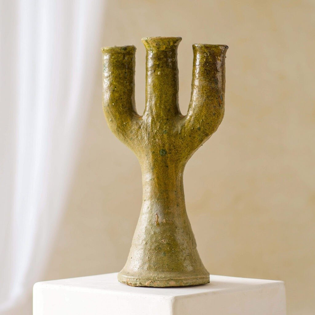 A rustic brown ceramic vase with three cylindrical openings, reminiscent of a candelabra, sits elegantly on a white pedestal against a serene neutral background.