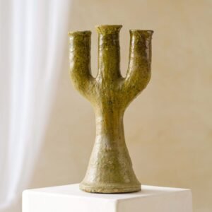 A rustic brown ceramic vase with three cylindrical openings, reminiscent of a candelabra, sits elegantly on a white pedestal against a serene neutral background.