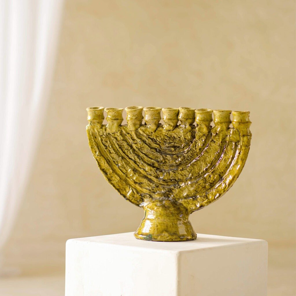 A ceramic menorah with a textured yellow surface is elegantly displayed on a white pedestal against a soft beige backdrop.
