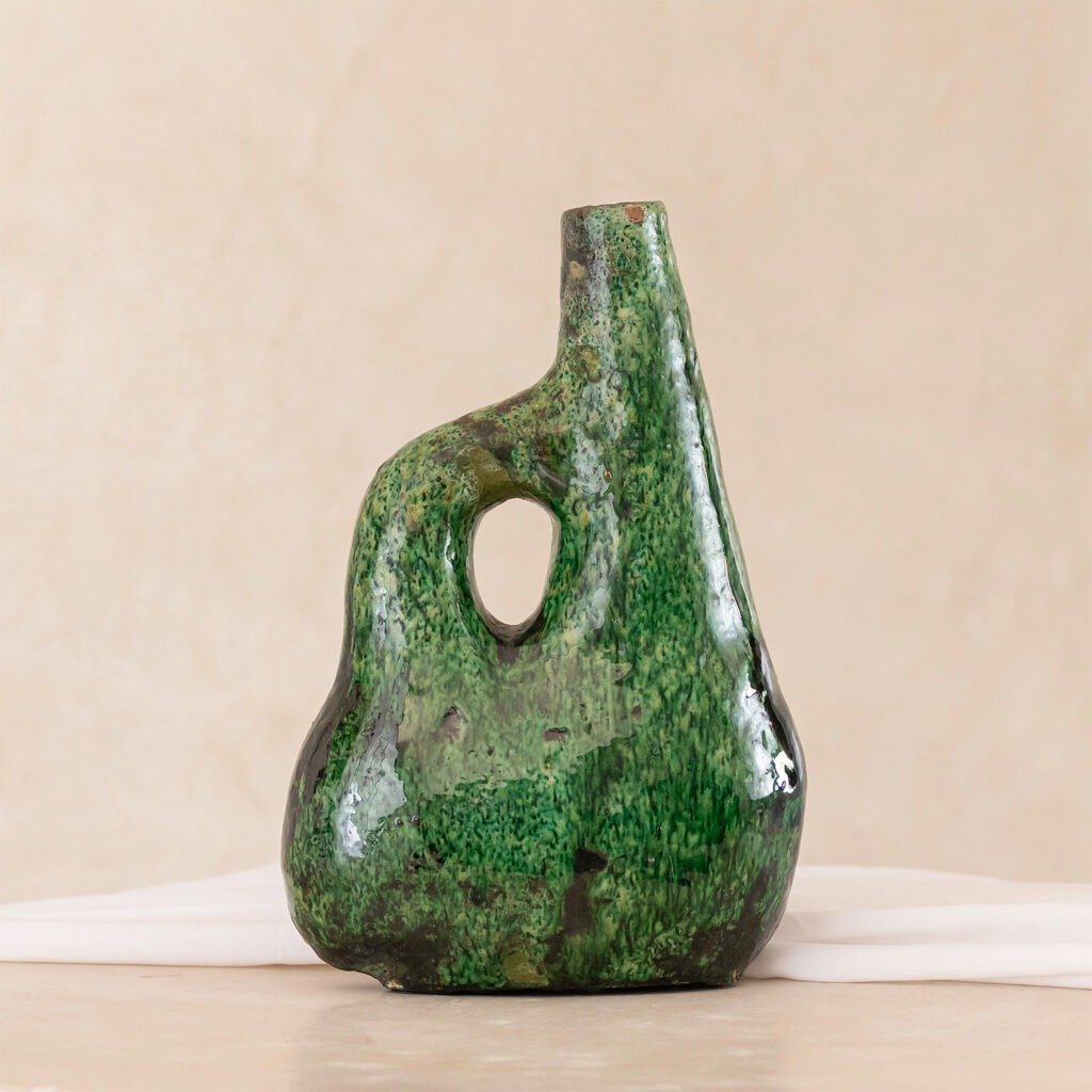 A uniquely shaped green ceramic vase with an irregular, rounded design and a hole near the top, set elegantly on a white surface.