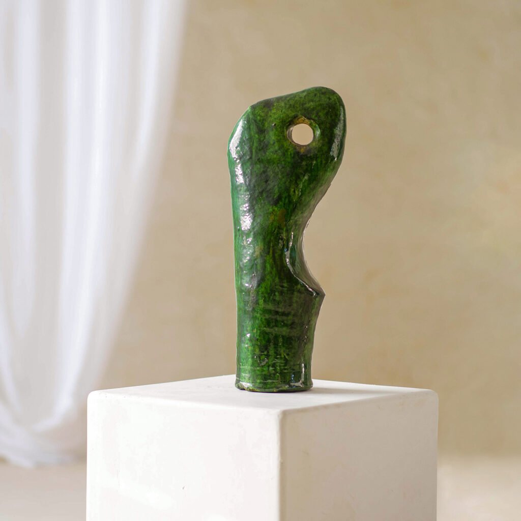 This green abstract sculpture, reminiscent of a serene wave, features a unique hole in its tall, curved form. Elegantly showcased on a white pedestal, it stands out against the neutral backdrop.