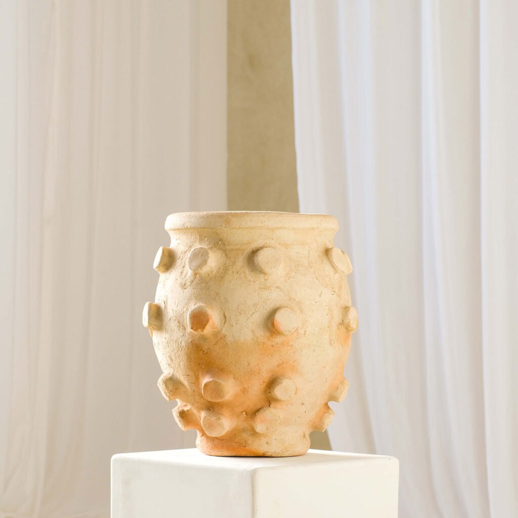 A beige, textured ceramic pot with protrusions is showcased on a white pedestal against an elegant backdrop of white curtains, exuding a timeless appeal.