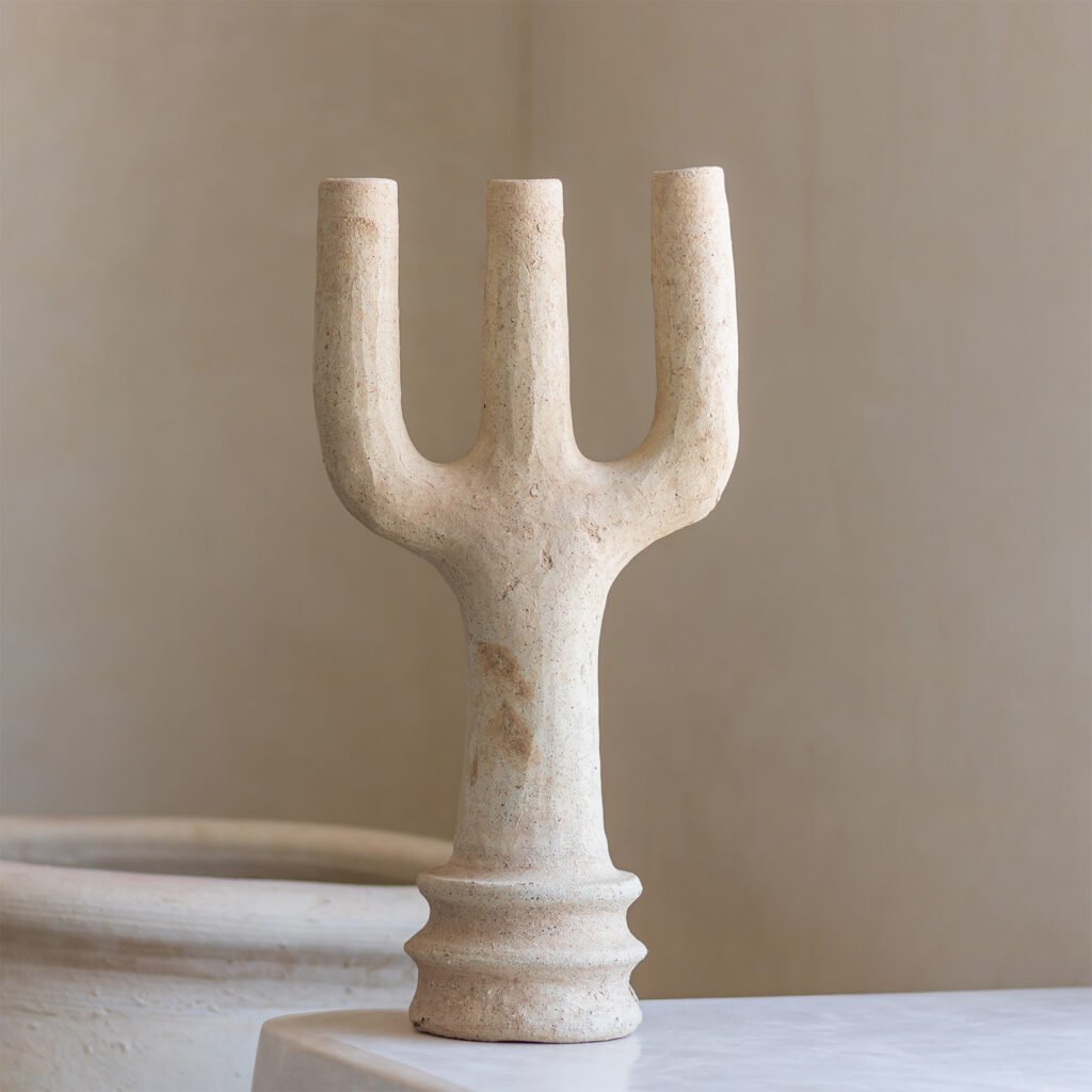 A beige, three-pronged abstract sculpture with a textured surface rests elegantly on a light-colored table, set against a minimalist backdrop.