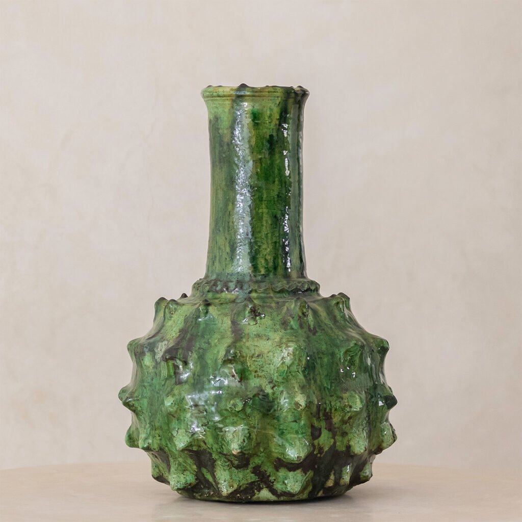 This green ceramic vase features a textured, bulbous base and a tall, narrow neck. Its surface offers an uneven, glossy finish, adding a unique touch to any space.
