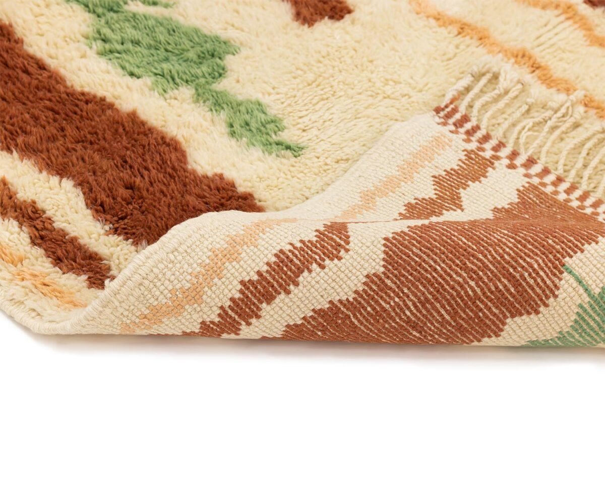 Close-up of a luxurious, hand-knotted multi-colored woven rug with earthy tones of brown, green, and cream. The underside reveals a different pattern featuring stripes.