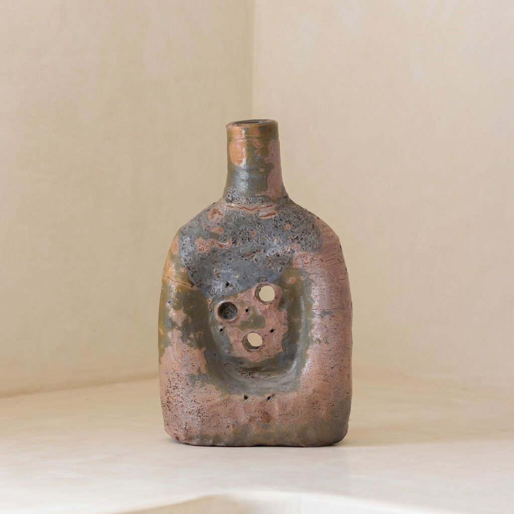 A rustic, rectangular ceramic vase with a small neck sits on a surface. The weathered look of brown and green hues complements its circular cutouts at the center, adding charm to any space. Consider pairing it with complementary colors or patterns for an inviting, cohesive decor.