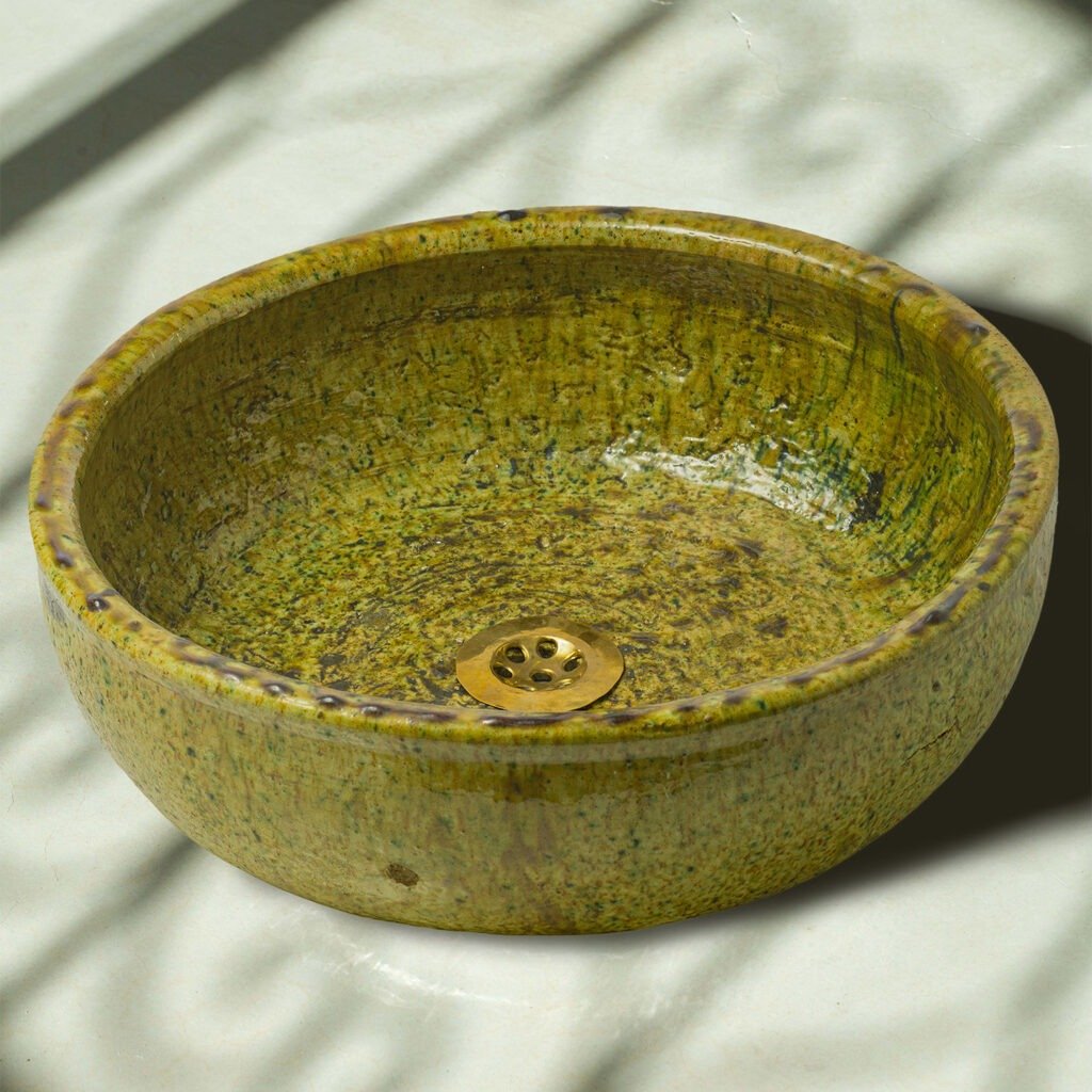 A round, textured, green ceramic basin with a gold drain sits prominently on a white surface, casting intriguing shadows. The unique blend of colors adds an artistic touch to any interior setting.
