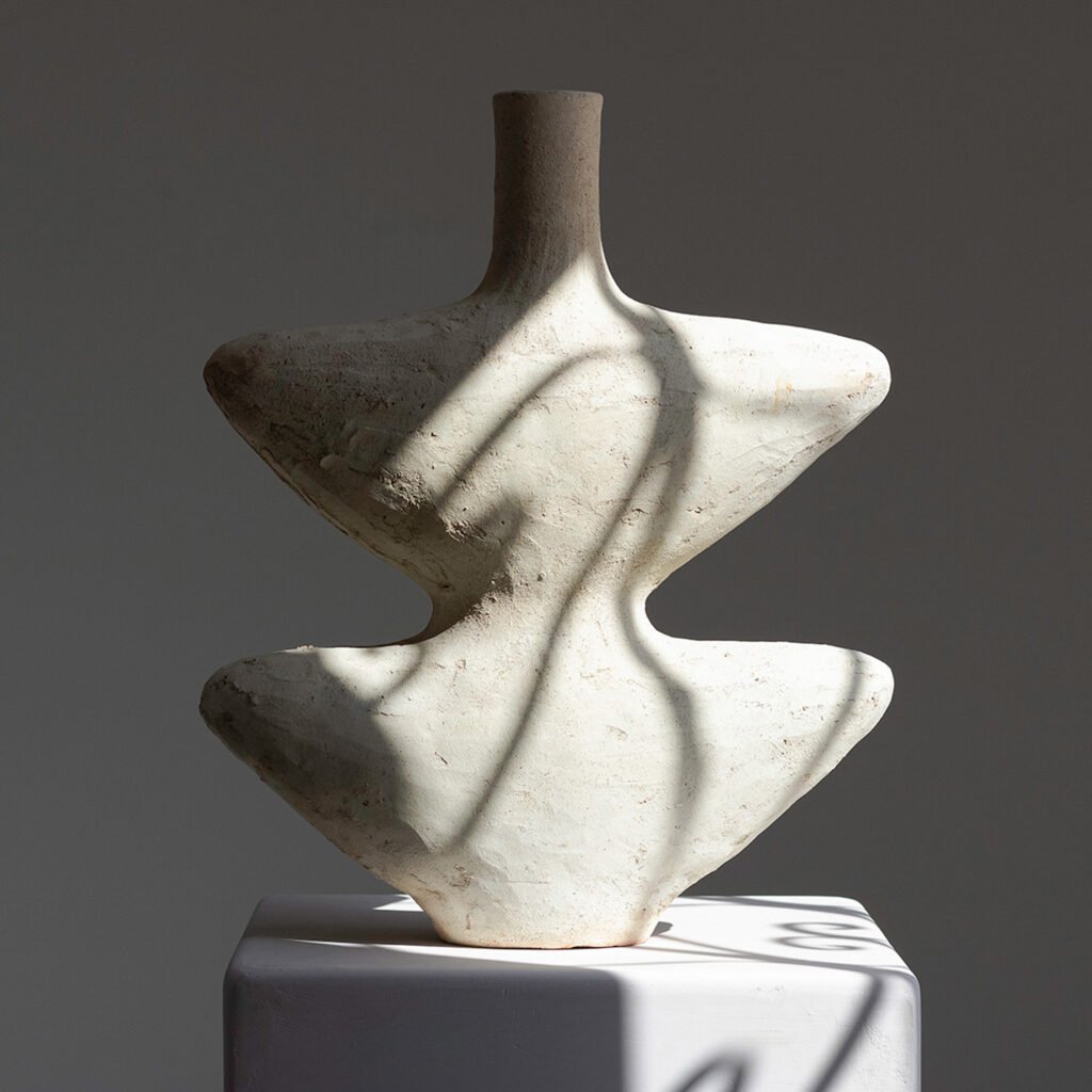 A textured, abstract ceramic sculpture with a narrow neck stands on a pedestal, casting soft shadows. The piece boasts an organic, asymmetrical form that evokes a sense of timeless elegance under diffused lighting.